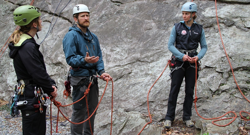 Three people who appear to be instructors wearing safety gear and stand near a rock wall. 
