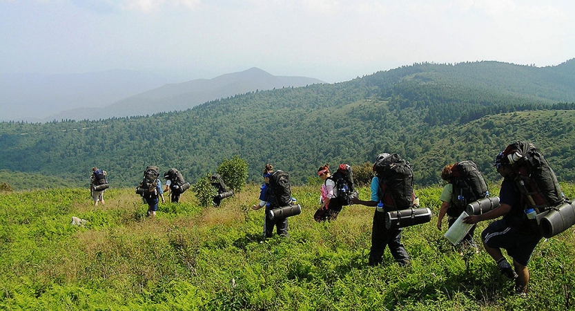 A group of people wearing backpacks hike along a green open space. In the back ground, the blue ridge mountains stretch into the mist. 