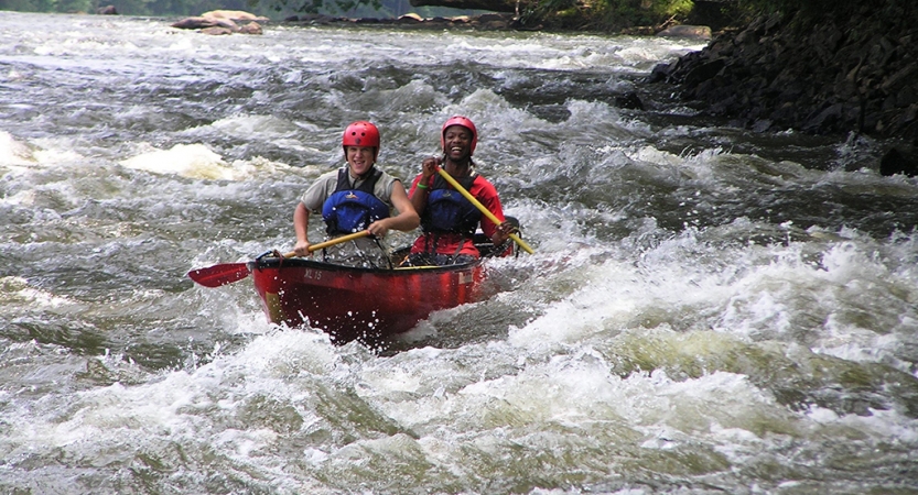 Two people wearing safety gear paddle a canoe through whitewater 
