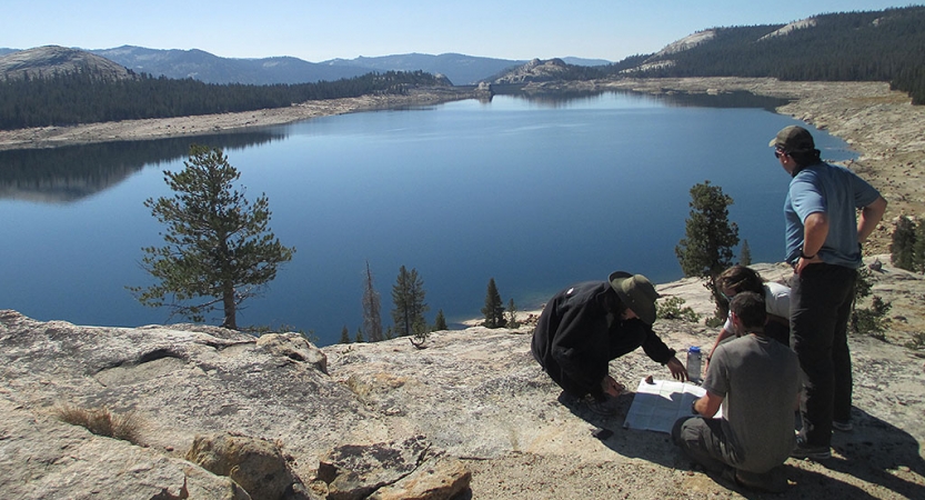 Three people sit on a rock near a body of water. 