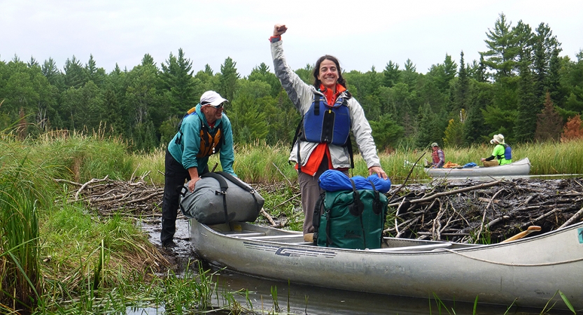 A person standing in shallow water beside a canoe raises their fist in celebration. There are other people in the background. . 