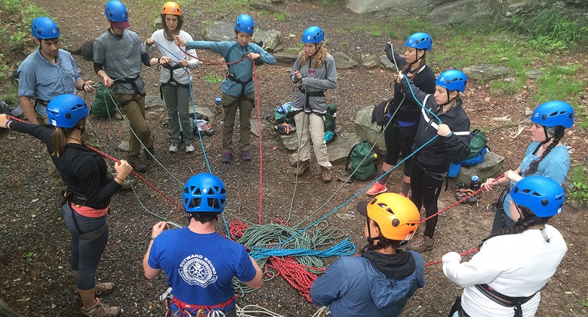 a group of people wearing safety gear stand in a circle on the ground. They appear to be engaged in an exercise, learning to tie climbing ropes. 