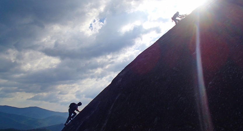 The silhouettes of two people climbing up a incline are illuminated as the sun moves behind them. 
