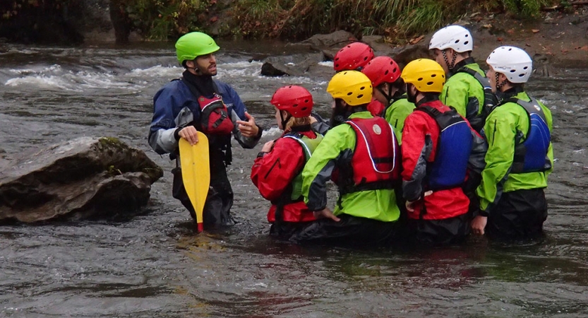 A group of people wearing helmets and lifejackets stand in waist-deep water listening to an instructor speak. 