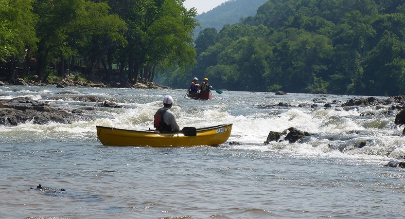 A person wearing safety gear sits in a yellow kayak floating near whitewater. The person in the kayak is watching another watercraft being paddled toward them. 