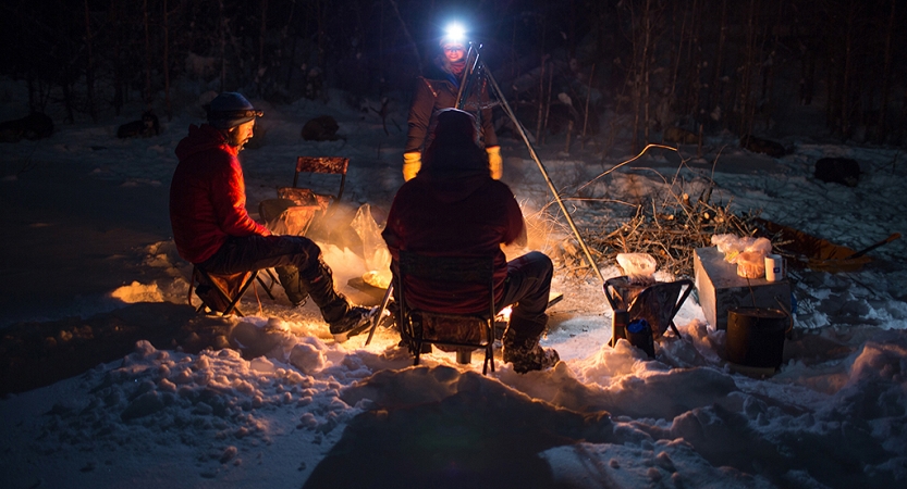 A group of people sit around a campfire at night in a snowy landscape. 