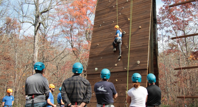 A person wearing safety gear is attached the ropes as they climb a man-made rock wall. Others wearing helmets stand on the ground and observe the climber. 