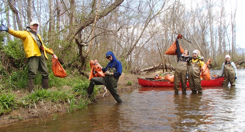 People stand in shallow water, holding bags of trash they collected from the area. 