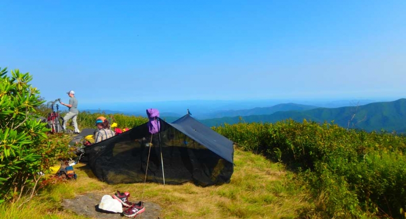 High above the vast blue ridge mountains, a tarp shelter rests on green grass. People are nearby and gear is scattered on the ground. 