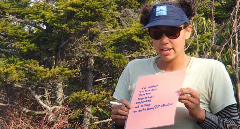 A person in a wooded area holds a sheet of paper with illegible writing on it.  