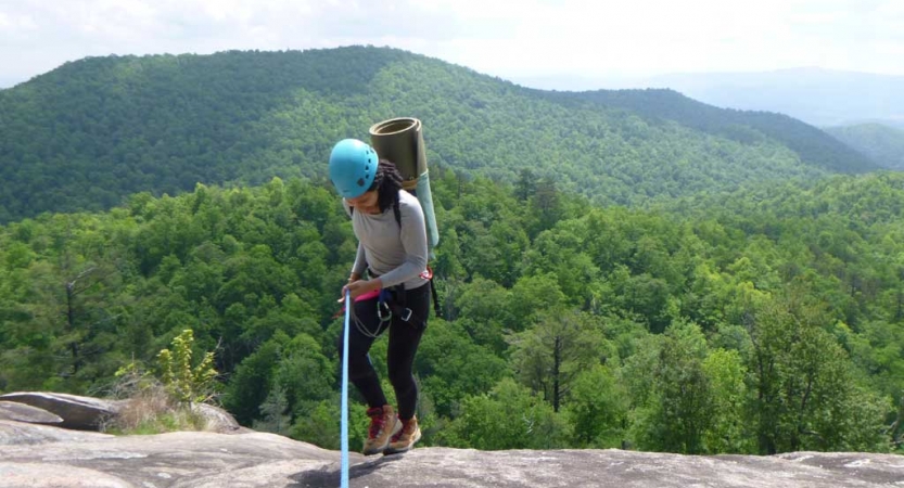 A person wearing safety gear is attached to a rope as they stand on a rock overlook, high above a mountainous green area below. 