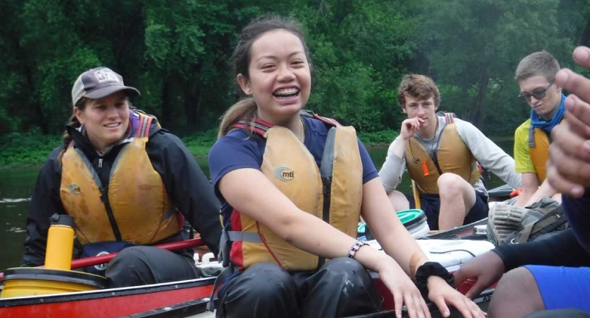 A group of students wearing lifejackets sit in canoes with a tree-lined shore in the background. 