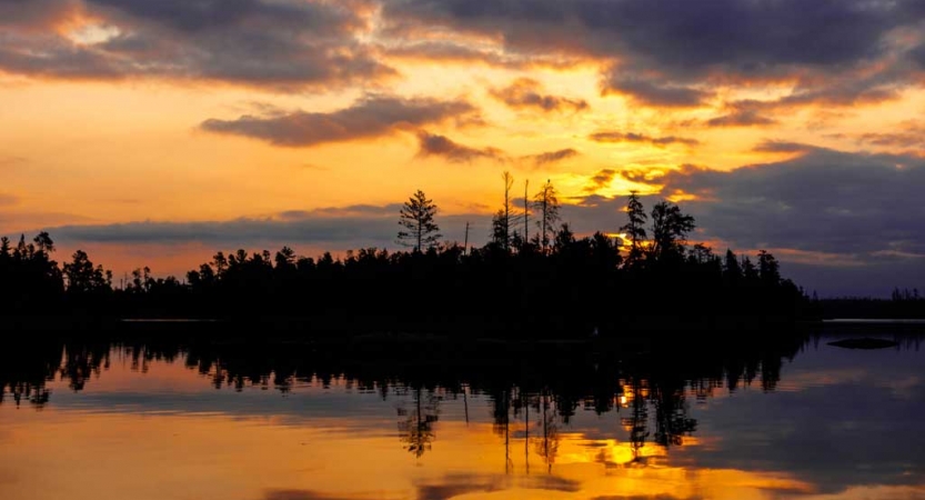 The sky appears in shades of yellow and orange as the sun either rises or sets behind a line of trees on the bank of a lake.