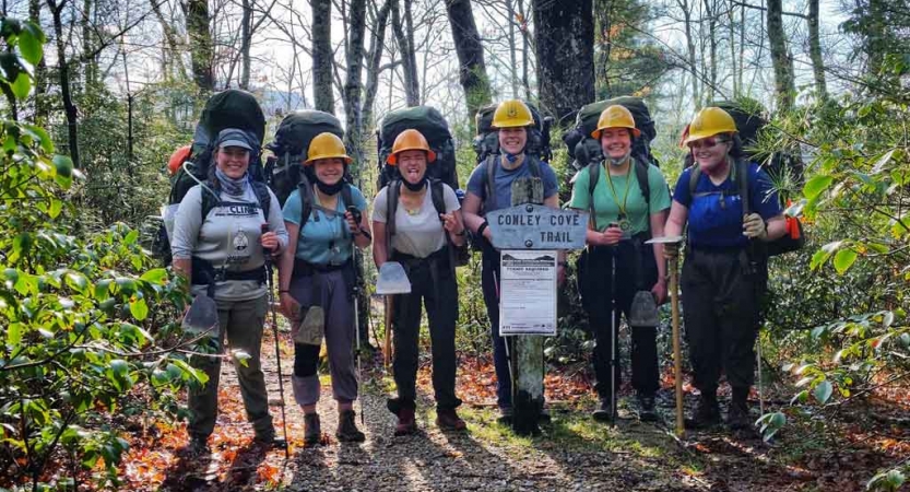 A group of people wearing backpacks and helmets stand in a line in front of a trail sign and smile.
