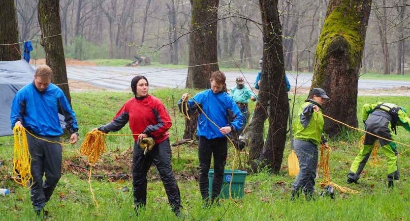 A group of people wearing rain gear stand in a green wooded area and appear to be working with climbing ropes. 