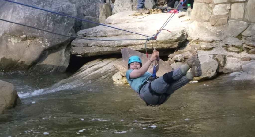 A person wearing safety gear is attached to a rope as they navigate across a river, demonstrating a Tyrolean traverse.