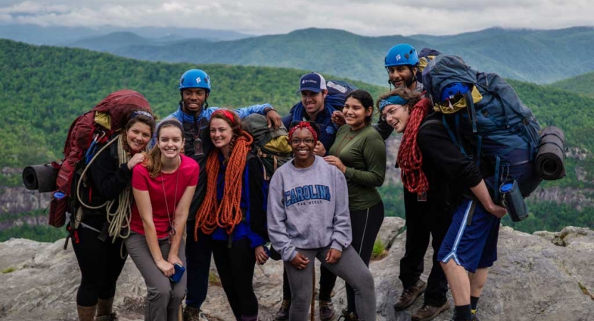 On a rocky overlook above the blue ridge mountains, a group of people pose for a photo. Many of them are holding various gear. 