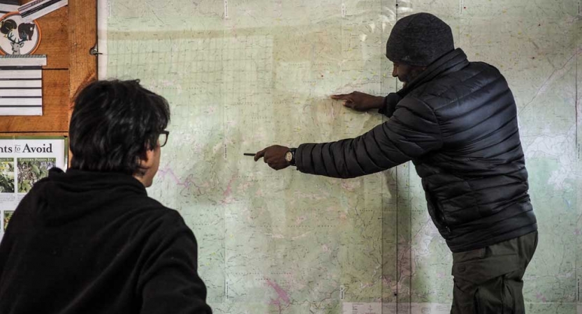 A person stands and points at a large hanging map while another person looks on.