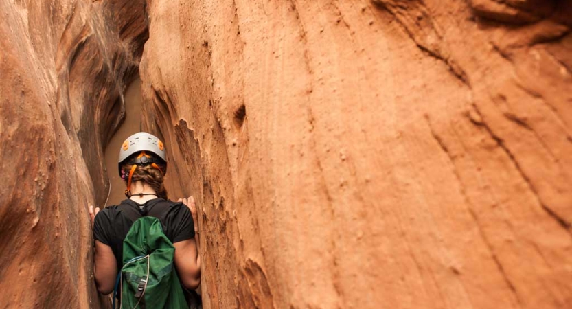 From behind, a person wearing a helmet moves through a red slot canyon 