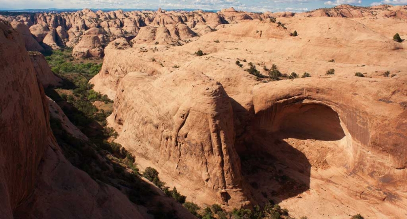 From above, a desert landscape marked by red canyon walls stretches into the distance. 