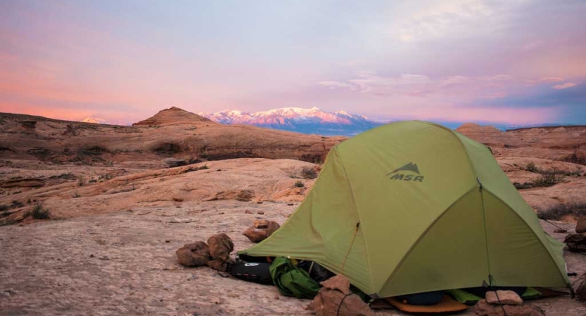 A tent rests in the sand with a mountain in the background. The sky is in shades of pink and purple. 