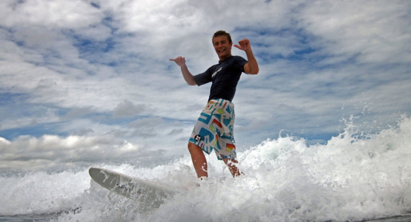 A person gives a hang loose sign while riding a wave on a surfboard 