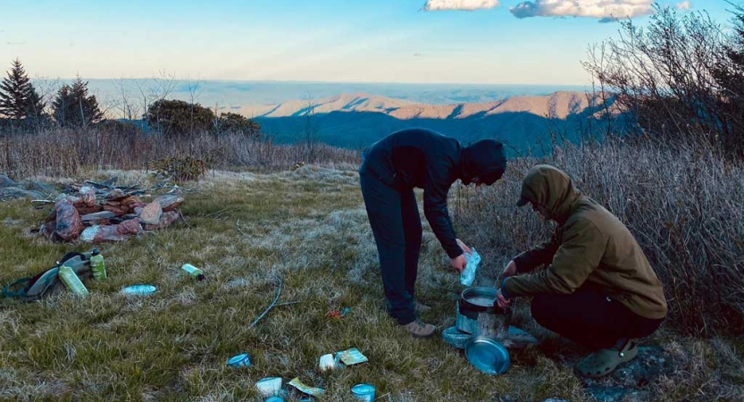 On a grassy overlook high above the outstretch blue ridge mountains, two people fill water bottles. Gear is scattered around on the ground nearby. 