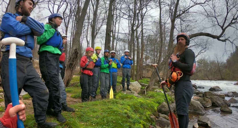 A group of people wearing safety gear stand on the bank of a river and listen to an instructor speak