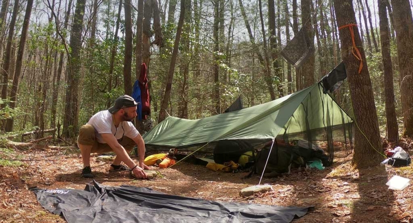 A person kneels beside a tarp shelter in a wooded area.