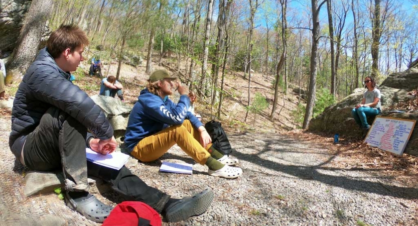 A group of people sit on the ground in a wooded area, listening to a person sitting near a whiteboard speak.