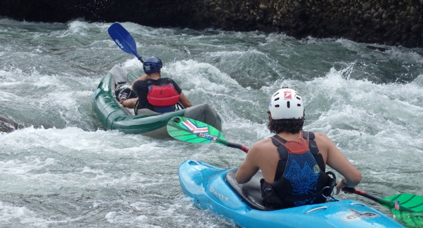 Two people wearing safety gear paddle two watercraft through whitewater. 