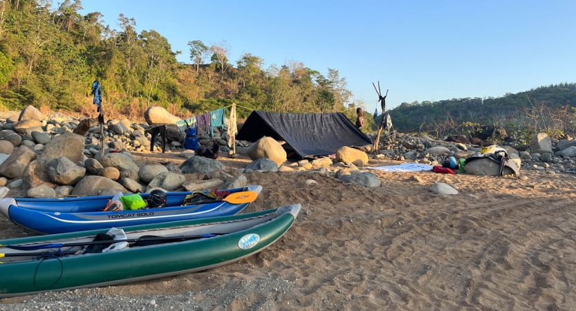 A tarp shelter and watercraft rest on the shore of a river near trees.