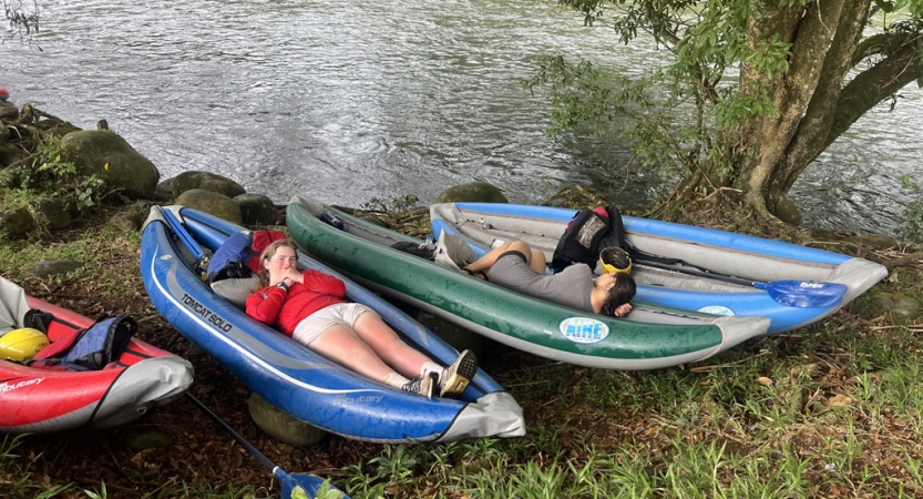 two people rest in beached watercraft on the shore of a river.