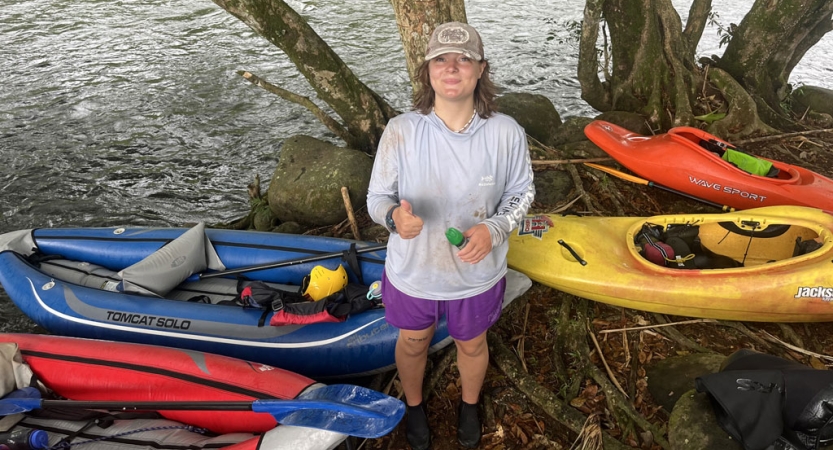 A person stands on the shore near beached watercraft and gives the camera a thumbs up.